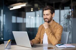 Mature businessman sitting at desk working on laptop with confidence and focus. Office worker in modern workspace showing dedication to work and thoughtful expression. photo