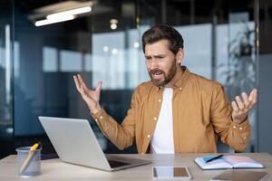 Mature businessman experiencing frustration at laptop in modern office. He gestures with raised hands, showing stress from remote work challenges or technical issues. Professional surroundings. photo