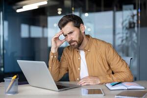 Stressed businessman in modern office environment, working on laptop. Man appears to be pondering difficult problem. Notebook and phone on desk emphasize technology and work pressure. photo