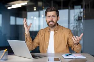 Frustrated businessman gestures at desk with laptop, notebook, phone, suggesting tech issue or confusion in office. Uncertainty and stress in workplace captured in moment. photo