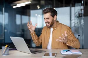 Stressed businessman at modern workspace reacting with frustration. Man sitting at desk with open laptop, notebook, phone, expressing anger or dissatisfaction in office setting. photo