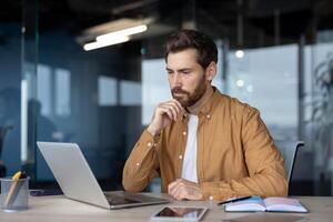 Mature businessman focused on laptop in office setting. Thoughtful expression indicates analysis and concentration. Notebook and phone on desk suggest professional work environment. photo