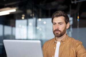 Man with beard using laptop in office. Reflects concentration, focus, productivity in modern workspace. Casual wear suggests informal but professional setting. photo