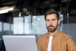Mature businessman with a confident expression using laptop in modern office. He exudes professionalism, focus, determination, dressed in casual attire, working on business tasks. photo