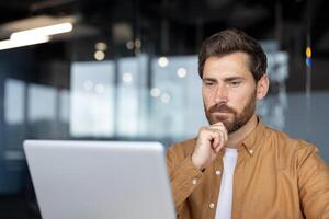 Mature businessman working on laptop in modern office, deep in thought. Man dressed in casual attire displays concentration and focus, reflecting on business strategies and solutions. photo
