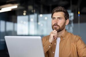 Man in casual shirt thinking while using laptop in modern office setting, reflecting on work ideas. Natural light filters through glass walls, creating focused atmosphere. photo
