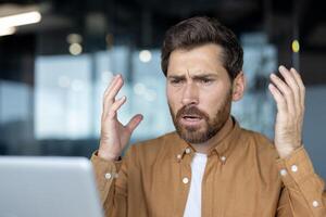 Mature businessman showing frustration in modern office, hands raised in disbelief. Professional expressing stress, likely due to unexpected challenge during important work task on computer. photo