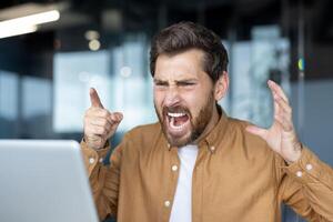 Frustrated businessman yelling at laptop during call. Intense anger and stress evident in facial expressions. Office setting hints at workplace conflict, emotion, frustration, and tension. photo
