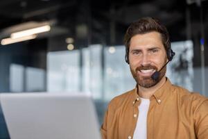 Mature businessman wearing headset working on laptop in modern office. Smiling professional involved in customer service or virtual meeting showcasing connectivity and teamwork. photo