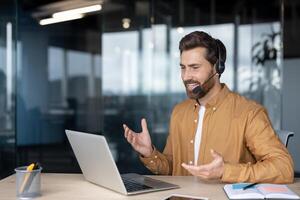 Mature businessman in office participating in call using laptop and headset. He appears engaged and communicative, gesturing during virtual meeting, highlighting remote work dynamics. photo