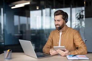 Mature businessman using laptop and headset while holding tablet. Engaged in online meeting in office setting. Demonstrates multitasking and communication skills in professional environment. photo