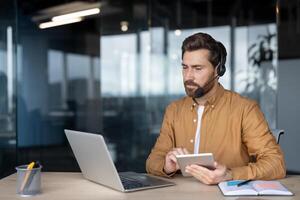 Business professional in office setting wears headset, uses laptop and tablet for remote work or virtual meeting. Concept of technology, communication, modern business photo