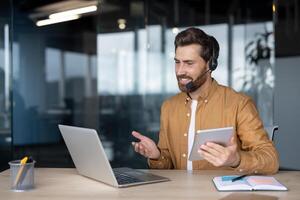Mature businessman in headset conducts online meeting using laptop and tablet. Professional setup at office desk for effective remote communication. Engaged in digital discussion on business topics. photo