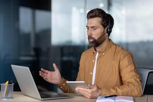 Mature businessman in office using laptop and tablet for conference, wearing headset. Engaged in communication, demonstrating multitasking skills. Professional setting enhances business atmosphere. photo