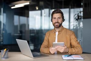 Smiling customer service representative wearing headset working remotely from modern office while holding tablet. Laptop on desk, showcasing professional technology use for communication. photo