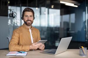 Confident mature businessman using tablet and laptop in modern office. Wearing headset for professional communication. Smiling, engaging, reflecting productive work atmosphere and technology use. photo