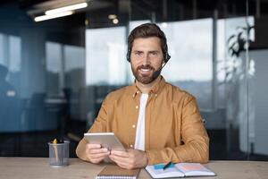Professional man wearing headset using tablet in modern office shows remote work technology. Confident expression, casual attire, seated with notebook and pen, signifies communication and teamwork. photo