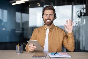 Mature businessman with headset using tablet, waving during virtual meeting in office. Confident and friendly expression, showcasing business communication and technology use. photo