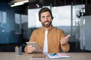 Business professional during call using tablet and headset, sitting in office. Gesture extends welcoming vibe. Conceptualizes modern communication, technology, workplace interaction. photo