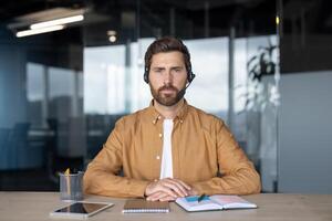 Professional man with headset at office desk using tablet for communication. Focused environment with modern workspace essentials like notebook and pen, embodying multitasking and digital interaction photo
