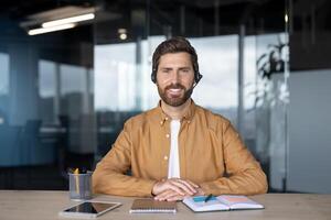 Mature businessman with headset sits confidently at office desk. Engages with digital technology such as notebook and tablet, portraying professionalism and productivity. photo