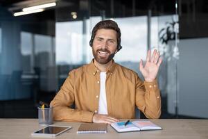 Smiling man in office wearing headset, engaged in call, waving hand. Friendly communication captured. At desk with notebook, tablet, and pen, conveying professional work environment. photo