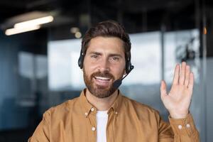 Confident man in office wears headset, smiling warmly while waving. Represents customer service, communication skills, professionalism. Ideal for business, support, tech, positive work environment. photo
