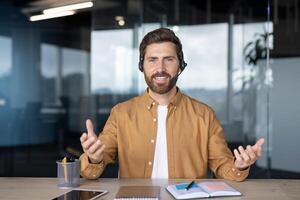 Smiling professional man with headset engaging in online meeting in modern office. Tablet and notebook on desk suggest active participation. Confidence, technology, remote work highlighted. photo