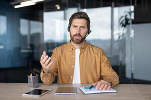 Mature business man with headset participating in online meeting. Expressive gestures enhance interaction. Desk features tablet, notebook, and pens, creating professional setting in modern office. photo