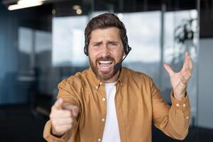 Frustrated customer service representative wearing headset in office expressing dissatisfaction through gestures. Pointing forward with annoyed facial expression suggests stress in workplace. photo