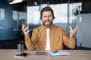 Businessman call confidently communicates while seated at office desk. Wearing headset, looking at camera, discussing with enthusiasm. Notepad and tablet add context of a professional setting. photo