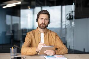 Professional man with headset in office taking notes during online meeting. Looking serious, sitting at desk with phone, notebook, pencil. Concept of modern communication, business. photo