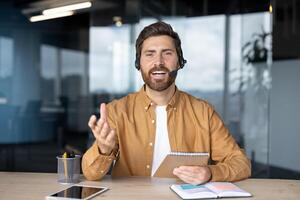 Confident businessman using headset, engaging in virtual meeting. Enthusiastic expression, holding notebook, gesturing during online discussion. Tablet and phone on desk enhance modern office setting photo