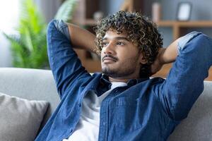 A young man relaxes on a couch with hands behind his head, contemplating in a home setting. The natural light enhances the serene ambiance. photo