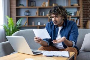A concerned man looks at a document with confusion while sitting at home. He seems stressed about bills or financial issues. photo