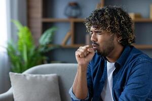 An afro-haired man, holding his fist to his mouth, coughs while seated on a sofa. It appears he may have a cold or similar illness. photo