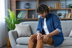 A man sitting on a couch clutches his knee in pain, indoors with shelves in the background. He appears to be experiencing discomfort. photo