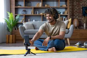 A man with headphones smiles while filming himself on a smartphone during a workout session at home, with gym equipment in the background. photo