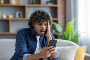A young man looks at his phone with a worried expression while sitting on a couch at home. photo