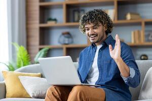 A smiling man waves during a call on his laptop while sitting on a comfortable sofa indoors. He is enjoying a friendly conversation. photo