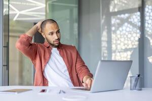 An African American man scratches his head looking thoughtfully at his laptop screen at a desk, facing some issue or idea. photo