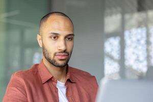 A focused, young man with a beard looks at a laptop screen, in a workspace with natural light filtering through the background. photo