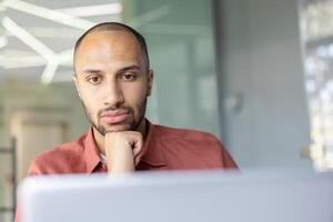 A pensive, dark-skinned man with a beard rests his chin on his hand in front of a laptop, lost in thought. photo
