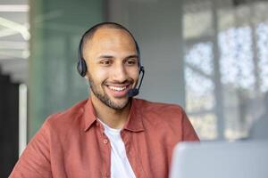 A smiling man wearing a headset appears to be in a customer service or call center environment, possibly assisting a client. photo