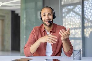 A smiling man wearing a headset gestures during a conference, sitting at a desk indoors, communicating. photo