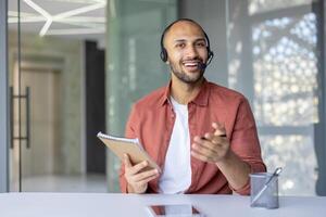 Webcam view, man with headset smiling and looking at camera holding notebook in hands. Company employee at online business conference meeting. photo