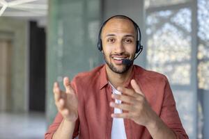 Webcam view, support worker with headset looking into camera, smiling, talking and consulting clients remotely. Man at workplace inside office photo