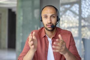 A man wearing a headset gestures while looking directly at the camera. He seems to be in a conference call, or an online meeting. photo