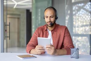A focused man wearing a headset looks at his tablet while working in an office setting. Natural lighting enhances the modern workspace. photo