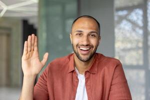 A happy man waves at the camera, smiling broadly and showing off his open palm. He's wearing a red-orange shirt. photo
